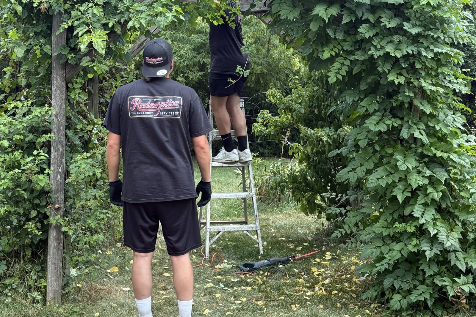 Two Redemption crew members in company shirts clearing an overgrown backyard arbor, one on a ladder cutting back vines while the other, in work gloves and a Redemption cap, watches from below.