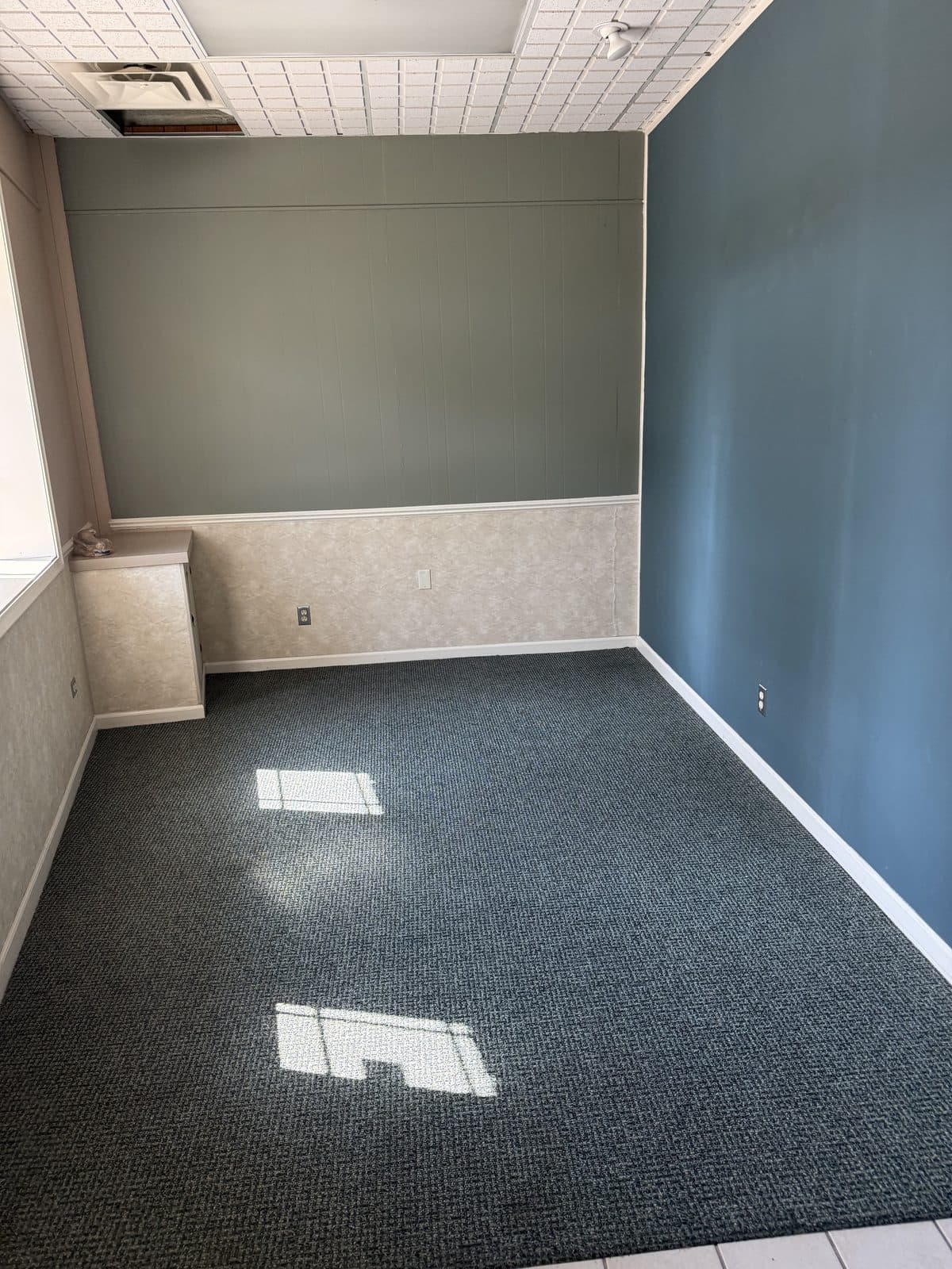 A carpeted private office with painted wall paneling and a suspended tile ceiling, before demolition.